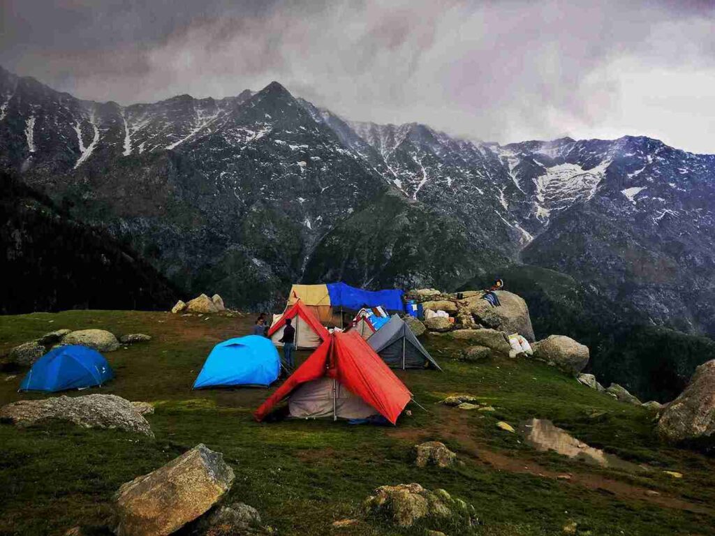 11 best places to visit in april in india Dharamshala Triund camping site with colorful tents, grassy ridge and Dhauladhar mountains under cloudy sky