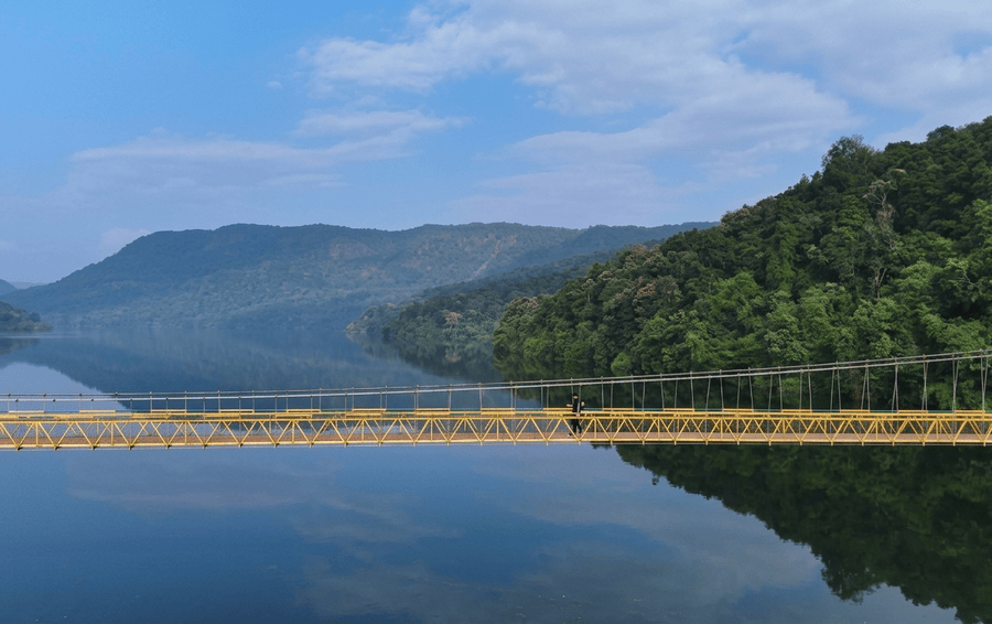 Wide panoramic view of a pedestrian hanging bridge spanning peaceful lake waters and lush mountain hillsides in Dandeli, highlighted among the best places to visit South India.