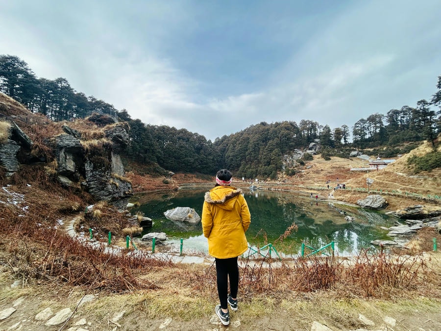 A traveler in a yellow jacket overlooking the serene Serolsar Lake near Jibhi, Himachal Pradesh, one of the 19 best places to visit in December 2026.