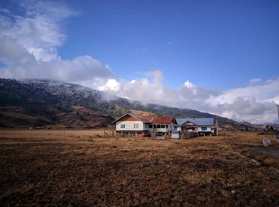 A scenic view of a wooden homestay in the Mechuka Valley, Arunachal Pradesh, under a blue sky with snow-capped hills, one of the 19 best places to visit in December 2026.