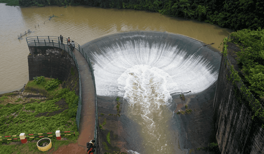 A high-resolution aerial view of the unique semi-circular spillway at Chiklihole Reservoir in Coorg, showing water rushing down the curved structure with onlookers watching from the adjacent walkway, featured as one of the 23 best places to visit in 2026.