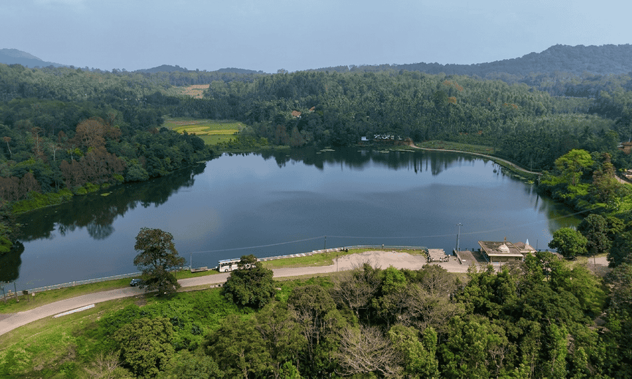 A high-resolution aerial view of Honnamana Kere Lake in Coorg, showing the serene water surrounded by dense green forests, with a small roadside temple complex in the foreground, featured as one of the 23 best places to visit in 2026.