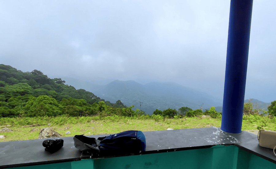 A high-resolution view of the rolling green landscape and distant misty valleys of Kabbe Hills in Coorg, as seen from a covered viewpoint with travel gear on the ledge and a blue pillar, featured as one of the 23 best places to visit in 2026.