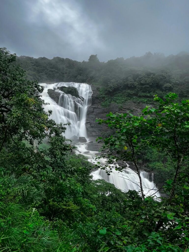A high-resolution view of the cascading Mallalli Falls, as seen in sumanth-hegde-zVq2LpBAbnY-unsplash.jpg, flowing down rock formations in Coorg. Dense green forests and foggy mountains under a cloudy sky frame the waterfall, featured as one of the 23 best places to visit in 2026.