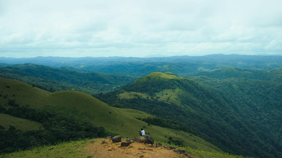 A traveler sitting on rocks overlooking the rolling green hills and misty valleys of Mandalpatti, featured as one of the 23 best places to visit in Coorg in 2026.