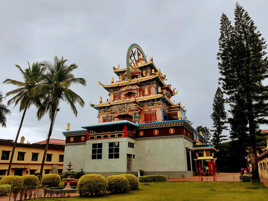 A high-resolution view of the ornate main temple of Namdroling Monastery in Coorg, as seen in tsewang-rigzin-sharpa-DCabHb8yh9g-unsplash.jpg, featuring its distinctive multi-tiered gold and red pagoda architecture and a dharma wheel on top, set within well-maintained gardens featuring a fountain and framed by a palm tree under an overcast sky, featured as one of the 23 best places to visit in 2026.