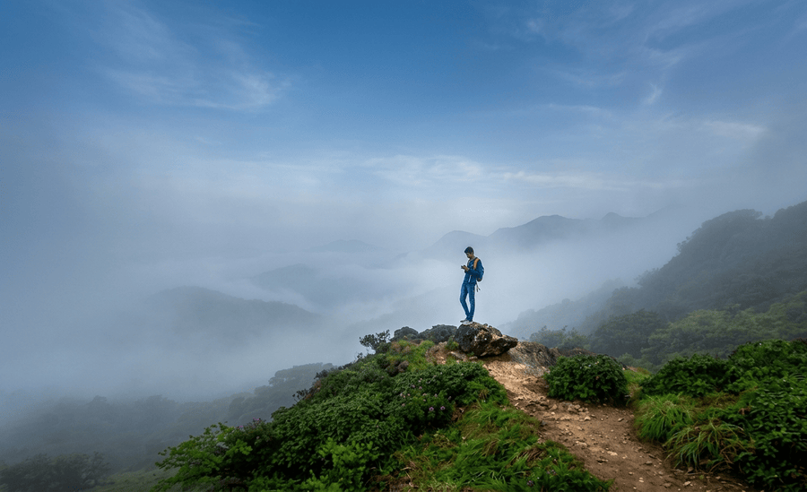 A lone trekker standing on a rocky outcrop at Tadiandamol Peak surrounded by misty hills and low-hanging clouds, featured as one of the 23 best places to visit in Coorg in 2026.
