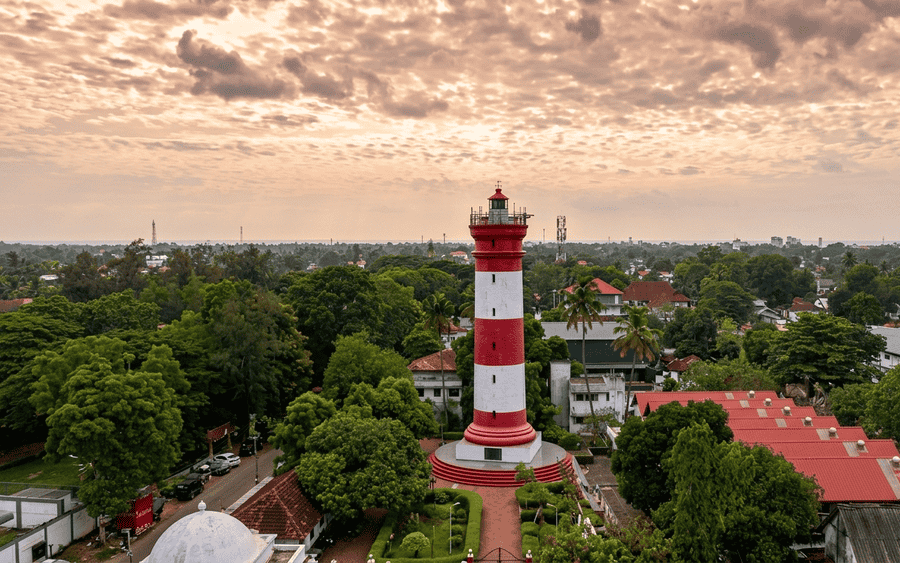 A high-quality, text-free aerial view of the historic red-and-white striped Alappuzha Lighthouse, one of the best places to visit in Alleppey, Kerala. The lighthouse stands tall among dense green trees under a dramatic cloudy sky, with local buildings and the water in the distance. All previous text and logos have been removed.