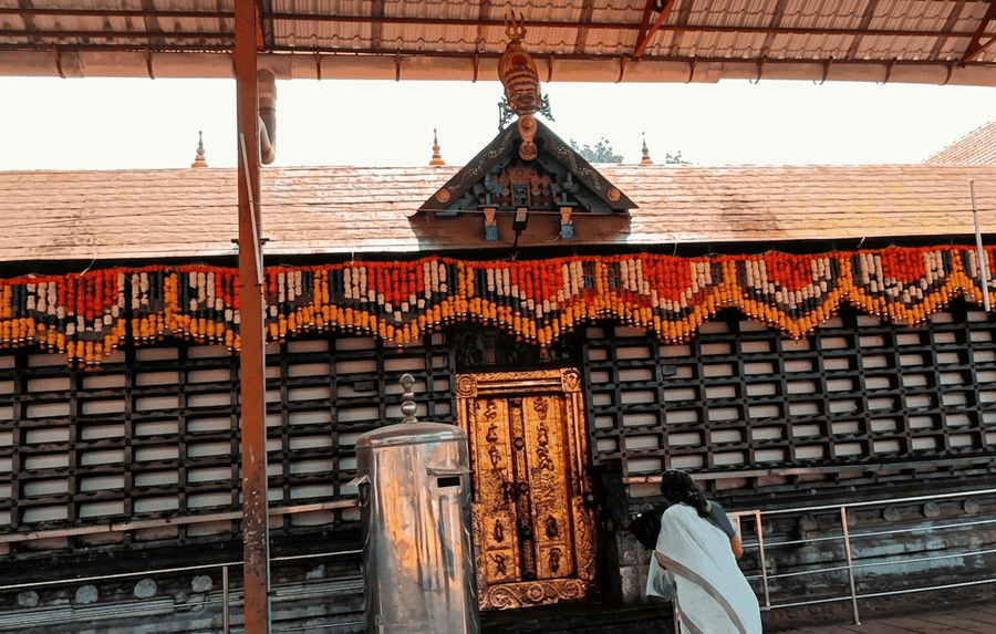 A detailed close-up photograph capturing the ornate, garland-adorned entrance of the Ambalappuzha Sree Krishna Temple, featuring a beautifully carved golden door, traditional Kerala temple architecture, and a prominent traditional deity mask figure on the gabled roof structure. It is considered one of the Best Places to Visit in Alleppey, Kerala.