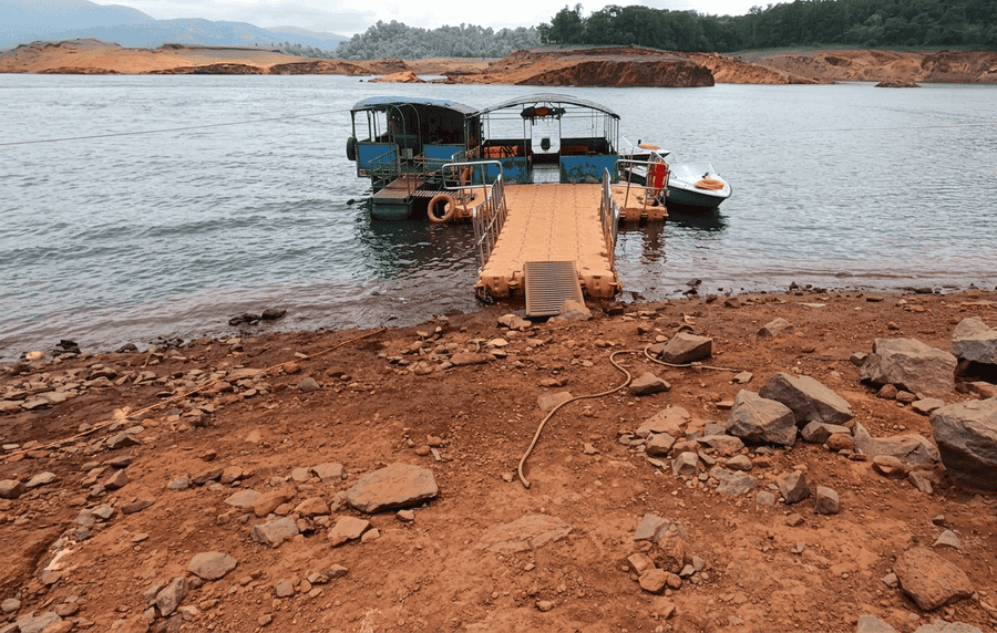A scenic view of the boat jetty at Banasura Sagar Dam, one of the best places to visit in Wayanad, surrounded by calm waters, red earth shorelines, and distant green hills under a cloudy sky