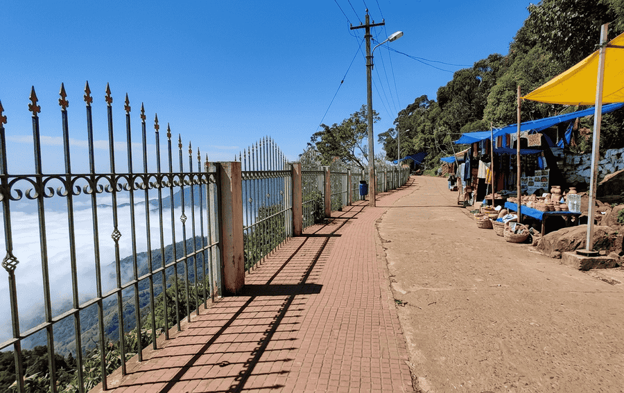 A wide-angle landscape photo of the scenic Coaker’s Walk path in Kodaikanal, showing a red-brick walkway, a decorative iron fence with a deep valley view below, and small stalls on the hillside, highlighted as one of the Best Places to Visit in Kodaikanal.