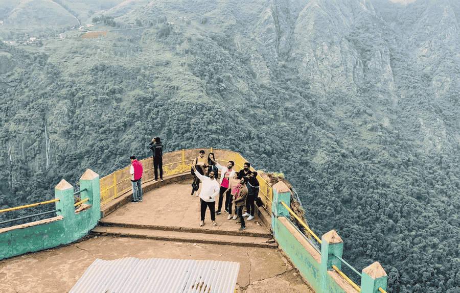 A high-quality, text-free scenic view of a group of tourists on the rocky outcrop of Dolphin's Nose, a premier landmark and one of the Best Places to Visit in Kodaikanal, overlooking the vast, forested Vattakanal valley and distant hills.