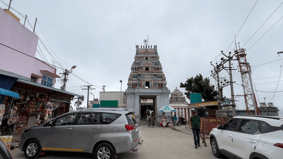 high-resolution, clear view of the intricately carved, multi-tiered South Indian style gopuram and main gate of the Kurinji Andavar Temple in Kodaikanal, under an overcast sky. The temple complex is vibrant and free of watermarks. In the foreground, a clean silver minivan on the left and a white SUV on the right frame the central street. Several individuals, including a man in a crisp green polo shirt, walk towards the temple gate. The scene captures the temple as a must-see landmark among the Best Places to Visit in Kodaikanal.