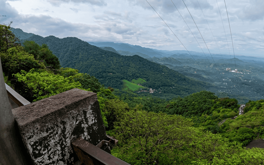 A panoramic landscape view from Lakkidi Viewpoint, one of the Best Places to Visit in Wayanad, showing lush green mountain ranges, a winding ghat road, and power lines under a cloudy sky.
