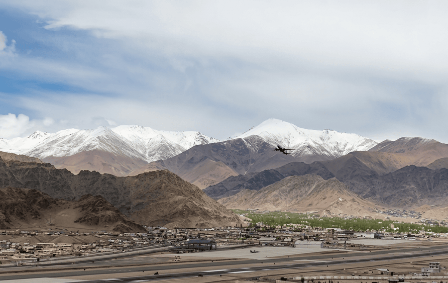 An expansive, high-resolution landscape photograph of Leh Local Sightseeing, a central element of any Leh Ladakh Itinerary, featuring the Leh airport, town, and valley nestled among massive, rocky mountains and distant snow-capped peaks. An aircraft is in flight above the valley floor.