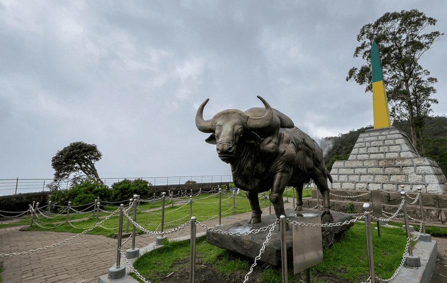 A scenic view of the large Indian Bison (Gaur) bronze statue and the blue-and-yellow-banded obelisk monument at Moir Point, a premier viewpoint and one of the Best Places to Visit in Kodaikanal.