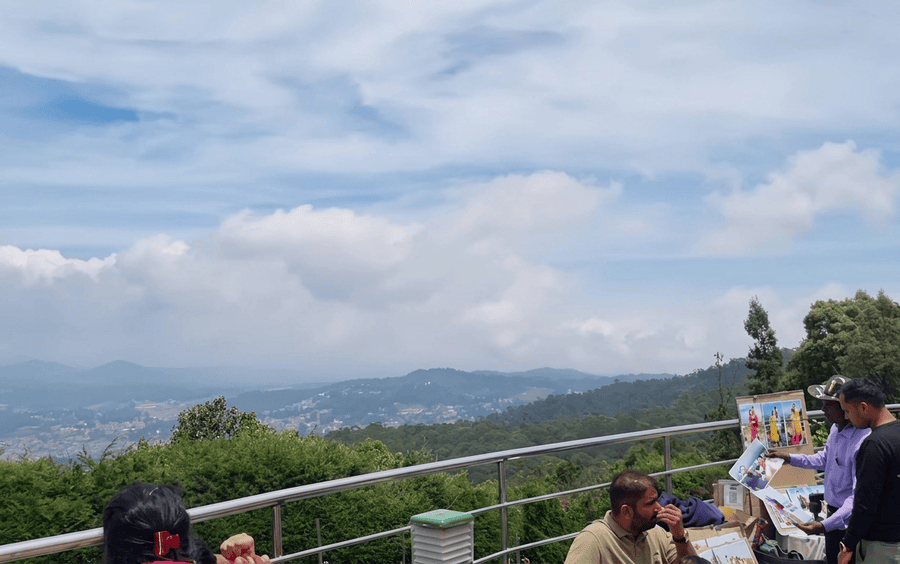 A panoramic mountain view from Doddabetta Peak, a must-visit viewpoint included in this Ooty Itinerary. Visitors on a platform overlook the valley, and a street photographer displays photos on the right.
