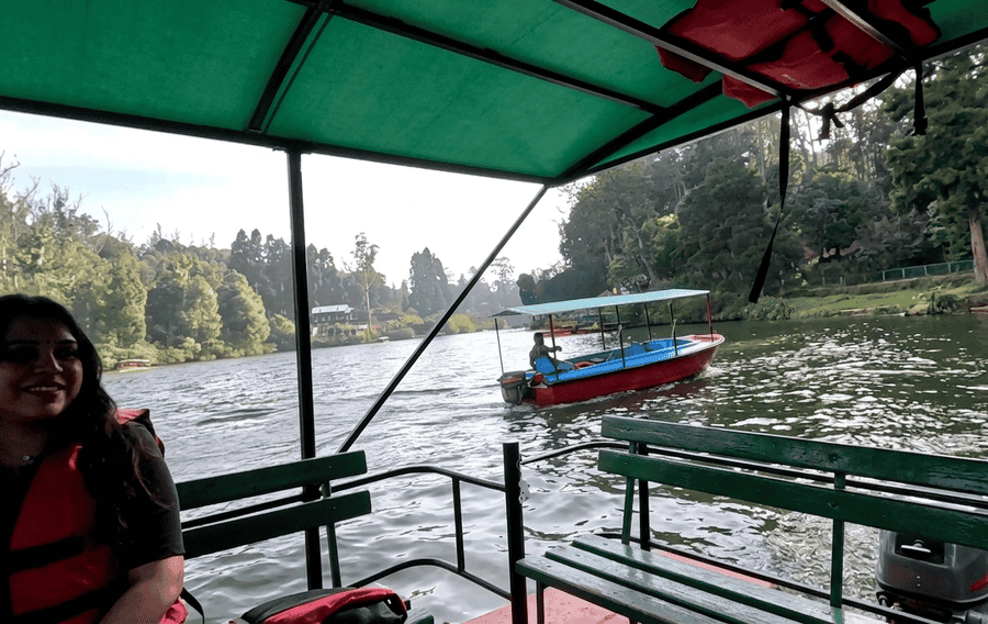 A view from inside a tourist boat on the tranquil Ooty Lake, surrounded by a lush forest of pine trees, a highlight on a classic ooty itinerary. A woman in a red life jacket smiles on the left, and another boat is visible on the water.