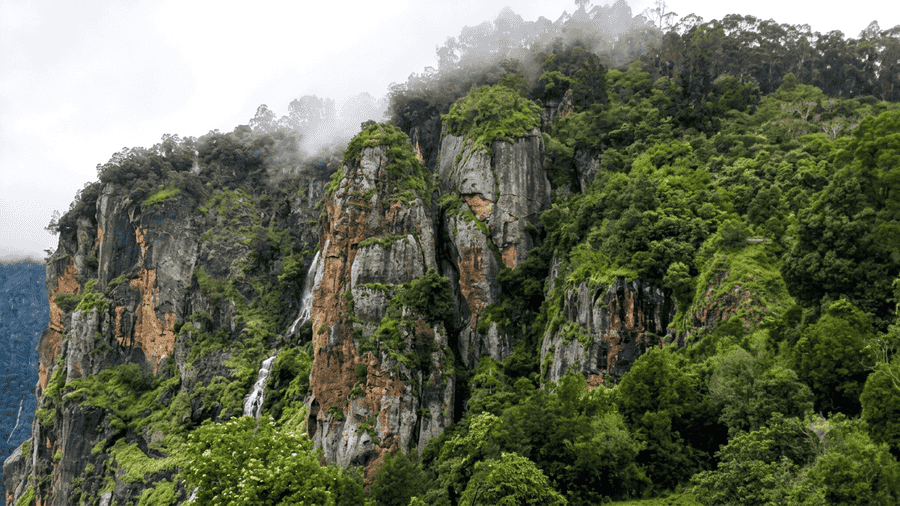 A high-quality, text-free scenic view of the towering Pillar Rocks formation and a cascading waterfall among the dense, misty forests, known as one of the Best Places to Visit in Kodaikanal, Tamil Nadu, India.