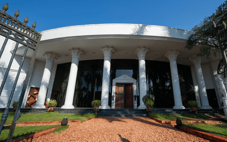 A wide-angle view of the elegant, classical facade of the Revi Karunakaran Museum, one of the **Best Places to Visit in Alleppey**, showcasing its grand white Corinthian columns, a curved portico, and large glass walls under a clear blue sky, framed by an ornate iron gate and flanked by classical statues.