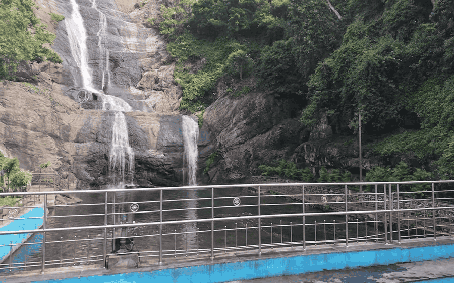 A high-quality, tourist-free photograph of the majestic Silver Cascade Falls, one of the best places to visit in Kodaikanal. The image shows multiple streams of water cascading down a large, tiered rock face, surrounded by dense green forest foliage. A detailed stainless steel safety railing frames the viewing area and lower pool. All crowds and logos have been removed
