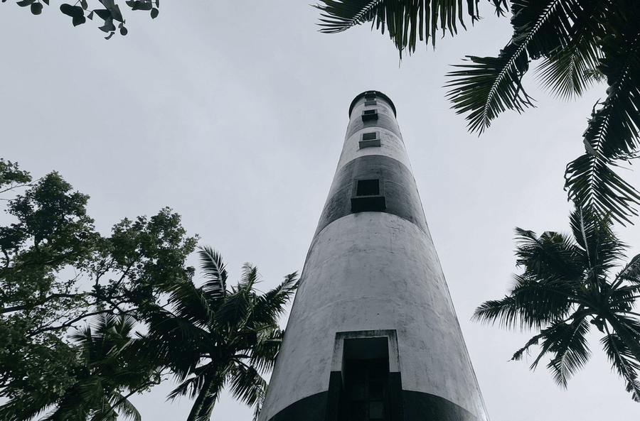 A low-angle view looking up at the black and white Anchuthengu Lighthouse framed by palm trees against a cloudy sky, a popular location among the best places to visit in Varkala.