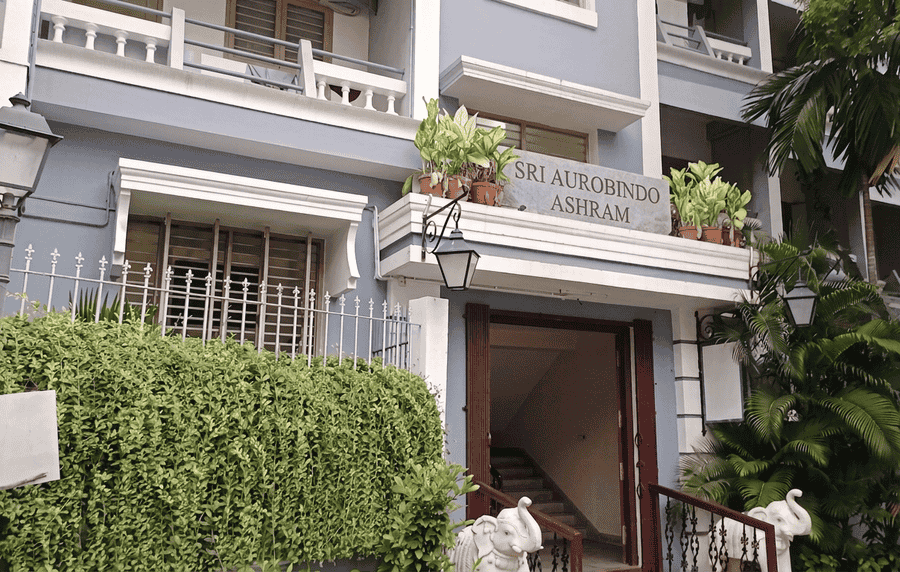 A photograph of the main facade and entrance to the Sri Aurobindo Ashram in Pondicherry, showcasing its colonial-style building, a sign with the ashram name, and flanking elephant statues. This ashram is universally listed as one of the best places to visit in Pondicherry for spiritual seekers and history buffs.