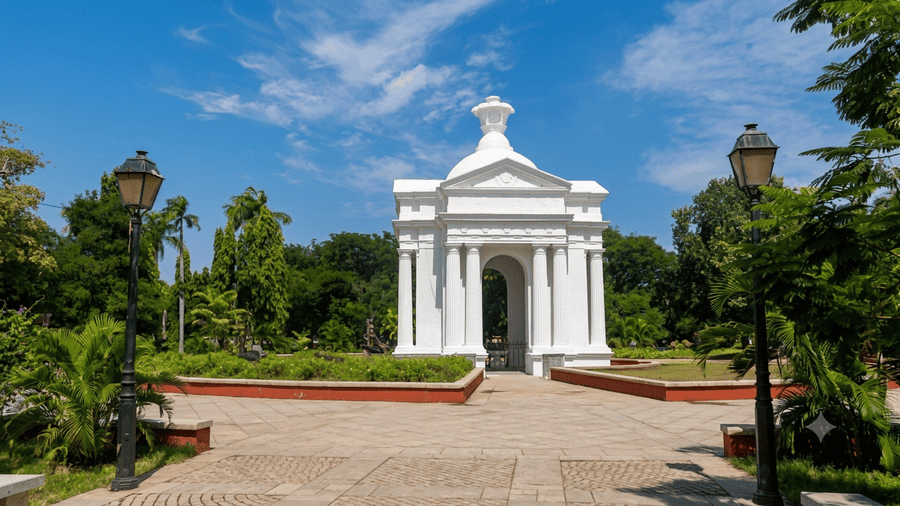 A bright daytime view of the Aayi Mandapam monument standing in the center of Bharathi Park, surrounded by green trees, paved walkways, and vintage lampposts. It is highlighted as one of the best places to visit Pondicherry for leisure and historic sightseeing.