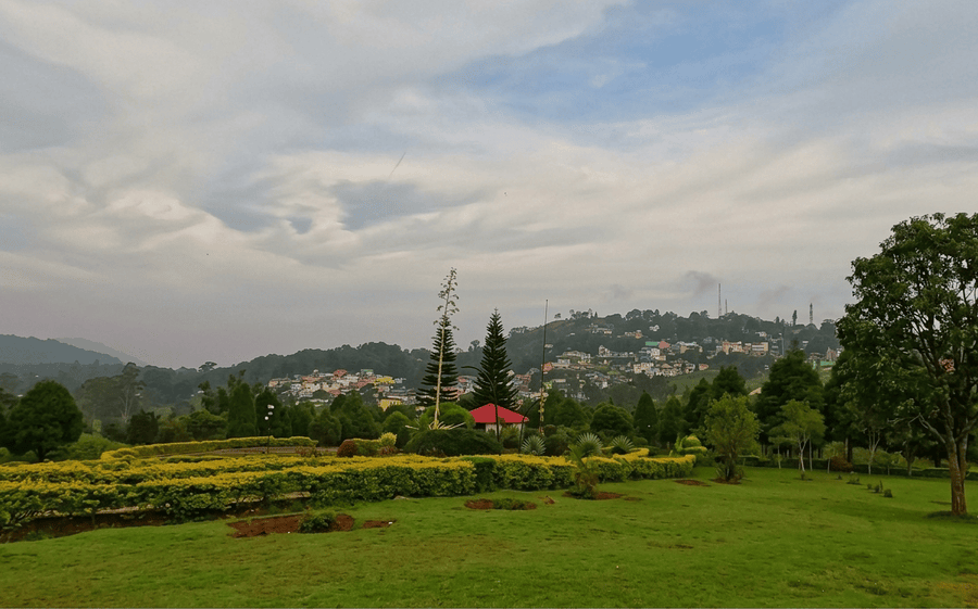 A wide, high-quality landscape photograph looking across the lush green lawns and hedges of the Botanical Garden towards a scenic hillside village, a top attraction among the best places to visit in Yercaud.