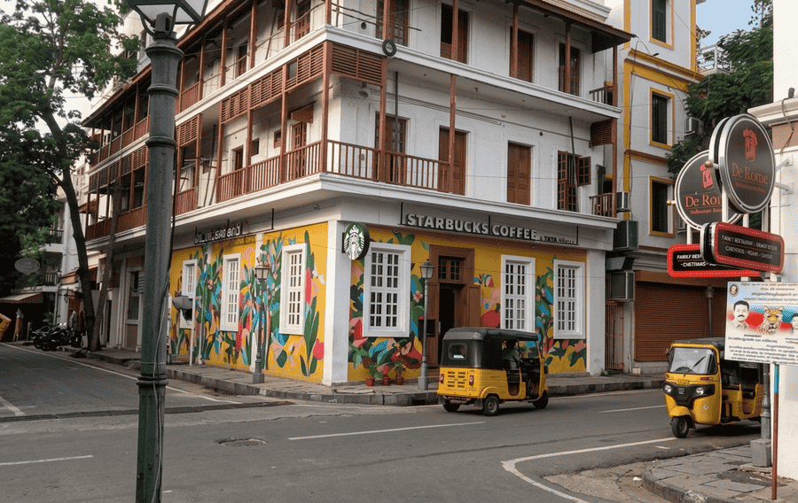 A colorful street in the French Quarter of Pondicherry, India, featuring a mustard-yellow Starbucks with a vibrant mural, a traditional yellow auto-rickshaw, and colonial architecture. It is known as one of the best places to visit Pondicherry for its heritage and unique blend of cultures