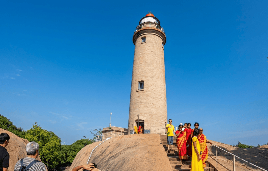 A vibrant, wide-angle photograph of the historic stone Mahabalipuram Lighthouse standing tall on a massive granite rock formation under a clear blue sky, showcasing why it is one of the best places to visit in Mahabalipuram, with a group of tourists walking in the foreground.