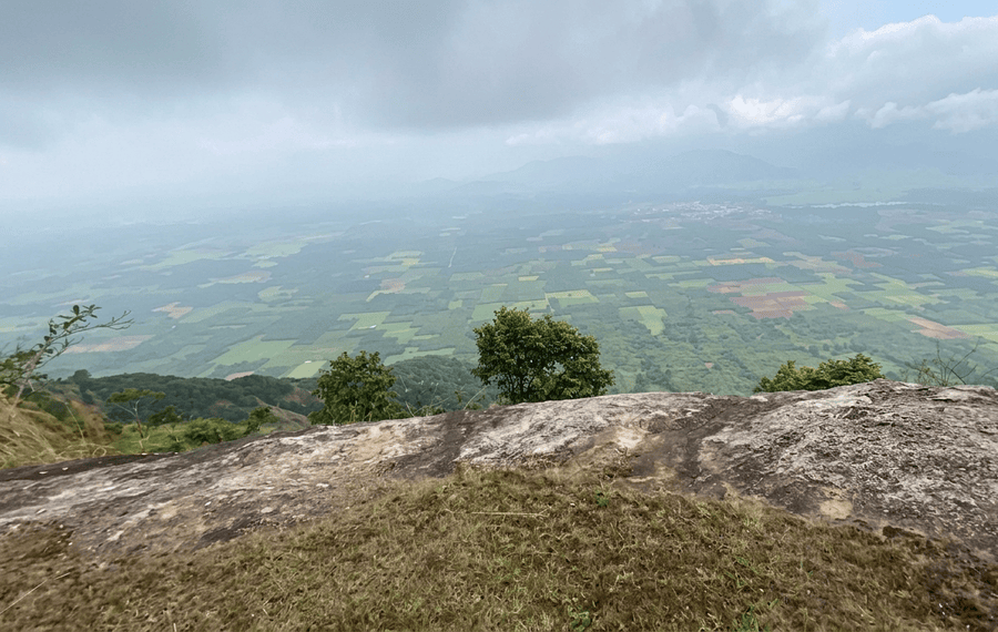 Standing on the rocky cliff at Pandikuzhi overlooking a patchwork of green agricultural fields under a cloudy sky, one of the best places to visit Thekkady.