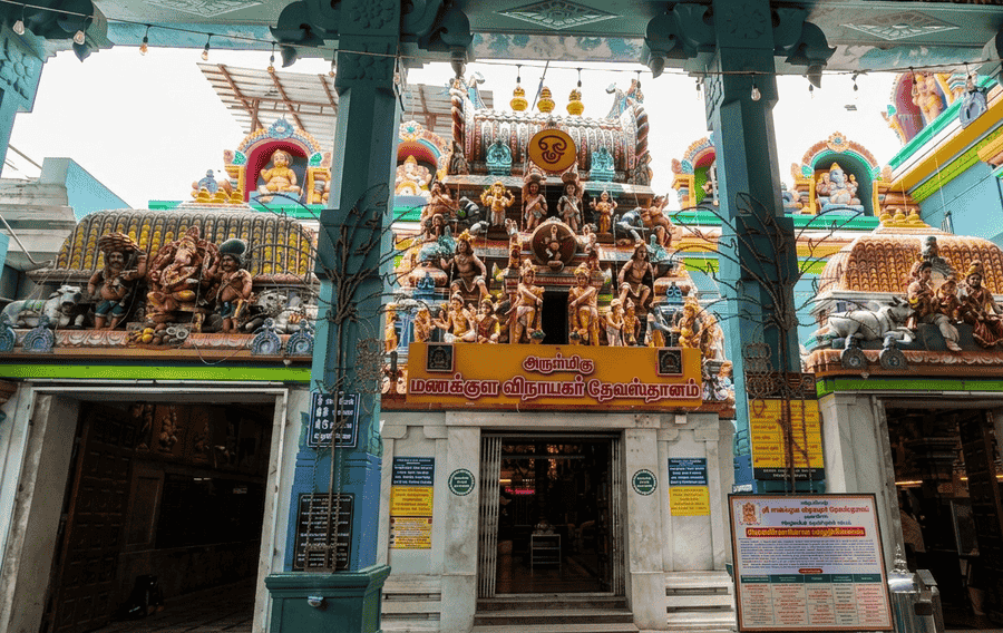 A colorful and highly decorative view of the Sri Manakula Vinayagar Temple in Pondicherry, showcasing its ornate facade with numerous Hindu sculptures, pillars, and a sign with Tamil text. This historic temple is globally recognized as one of the best places to visit Pondicherry for its cultural significance and unique Ganesha devotion.